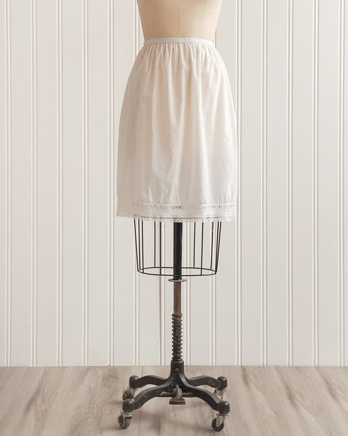 This image displays a vintage 1960s white nylon half slip, presented on a mannequin to highlight its fit and design. The half slip reaches approximately 23 inches in length and is tailored to accommodate a waist measurement of 12-14 inches, with the best fit for those with 36-inch hips.