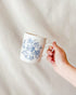 Hand holding a white mug with blue floral pattern on a neutral background